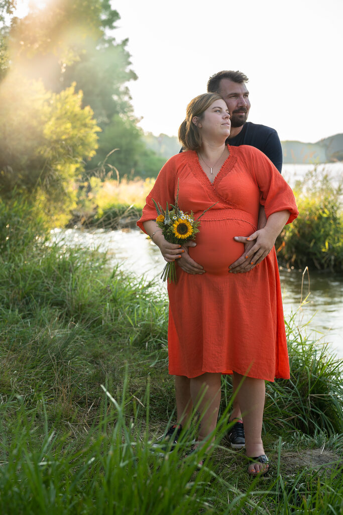 Mann umarmt Frau von hinten und beide haben ihre Hände am Bauch.