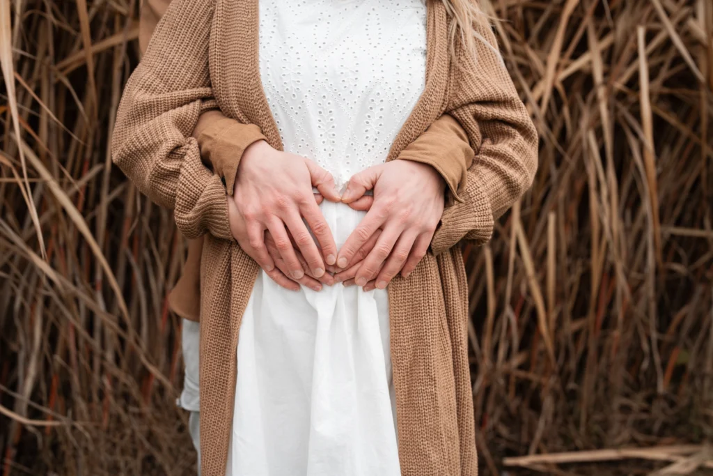 Nahaufnahme vom Babybauch. Mann umarmt Frau von hinten und beide Formen mit ihren Händen ein Herz am Bauch.