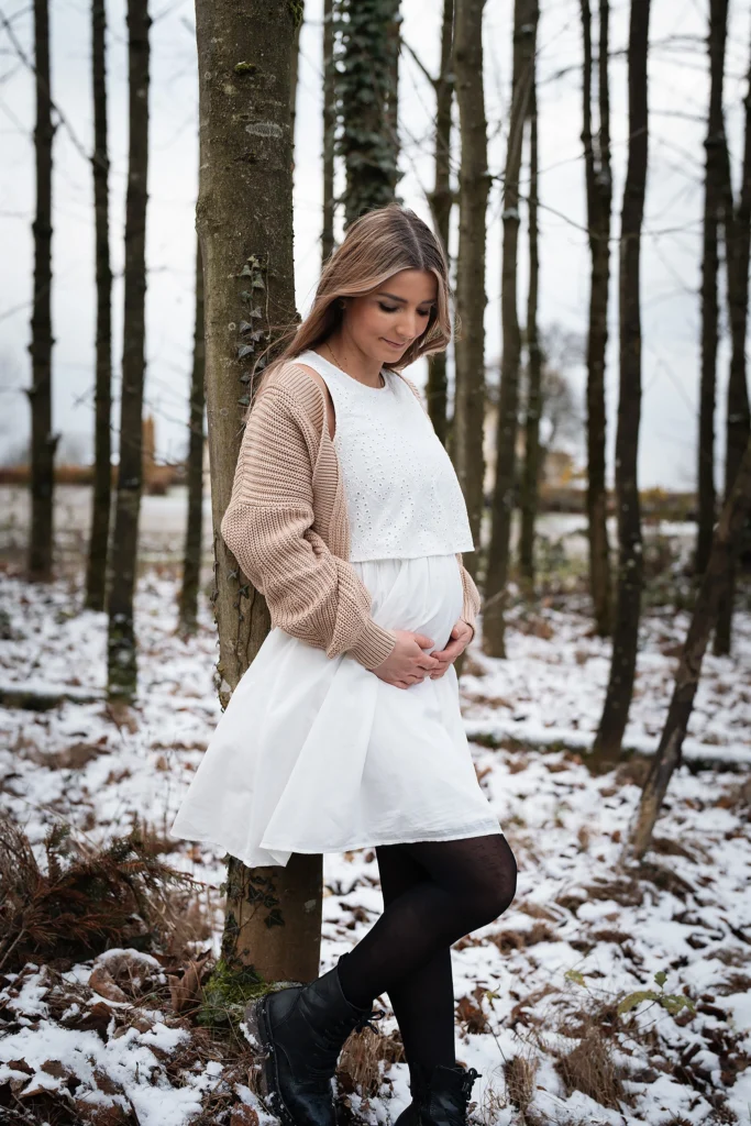 schwangere hübsche Frau in weißem Kleid mit beigen Cardigan angelehnt an einem Baum