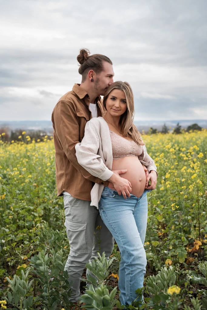 angehende Eltern in einem gelben Blumenfeld. Mann umarmt seine Frau von hinten, küsst sie am Hinterkopf und hat seine Hänge auf ihrem Babybauch.